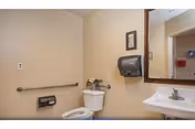 Small accessible bathroom with a toilet, grab bar, pedestal sink with mirror, and a wall-mounted paper towel dispenser.