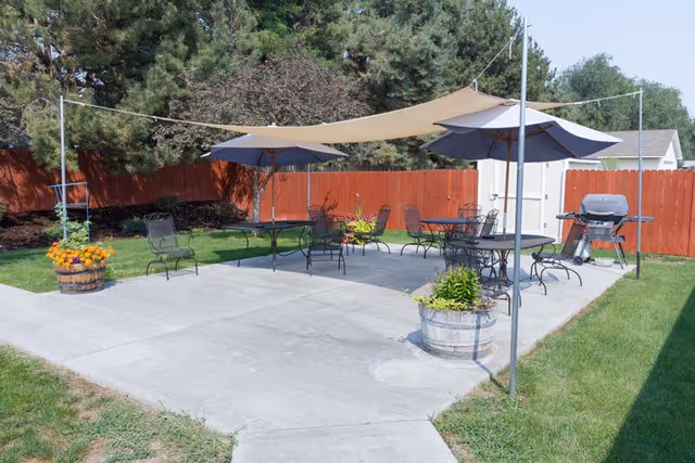 Outdoor patio area with several black metal tables and chairs under large umbrellas and a beige shade sail. There are potted plants and flowers around the patio, a red wooden fence in the background, and a barbecue grill near a white shed. Trees and green grass surround the patio.