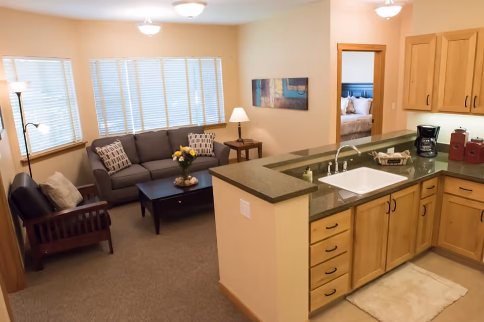 A cozy living room and kitchen area in a senior living facility. The living room has a gray sofa with patterned pillows, a wooden armchair with a cushion, a dark coffee table with a flower vase, and a floor lamp. The kitchen features wooden cabinets, a green countertop with a white sink, a coffee maker, and red canisters. A bedroom with a bed and pillows is visible through an open doorway.