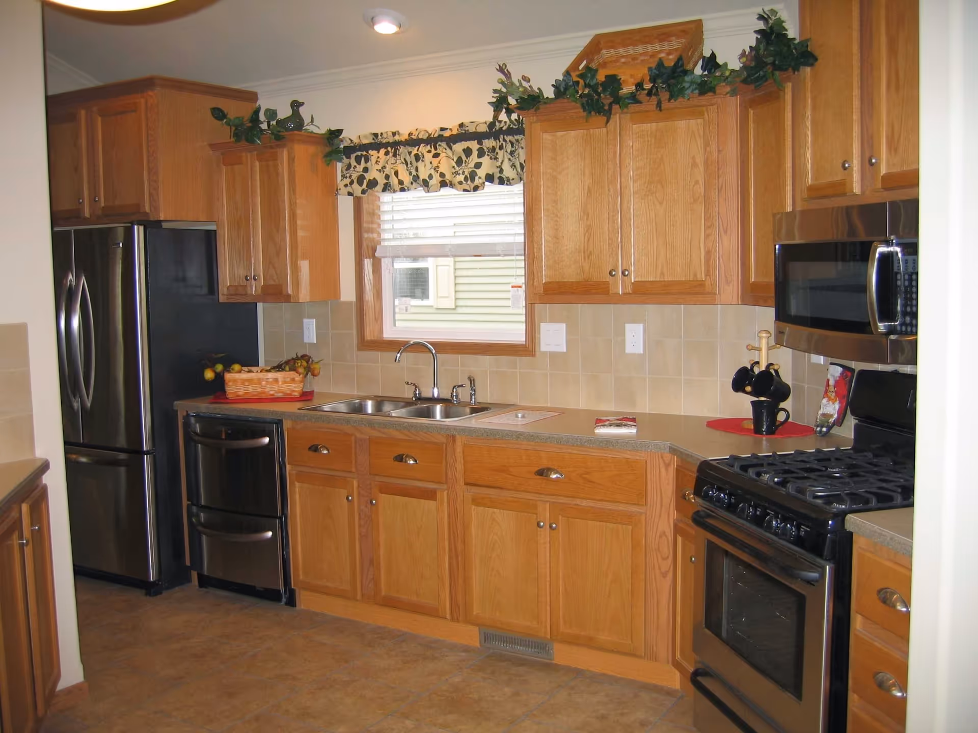 A kitchen with wooden cabinets, a double sink under a window with a floral valance, a stainless steel refrigerator, a black oven with a gas stove, and a microwave above it. There are decorative plants on top of the cabinets and a basket of fruit on the counter.