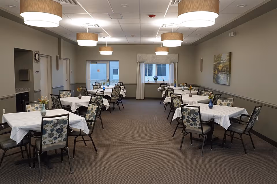 A dining room with several tables covered with white tablecloths, each surrounded by chairs with floral patterned upholstery. The room has beige walls, carpeted floor, large windows with curtains, and ceiling lights with round beige lampshades. Small flower vases are placed on each table.