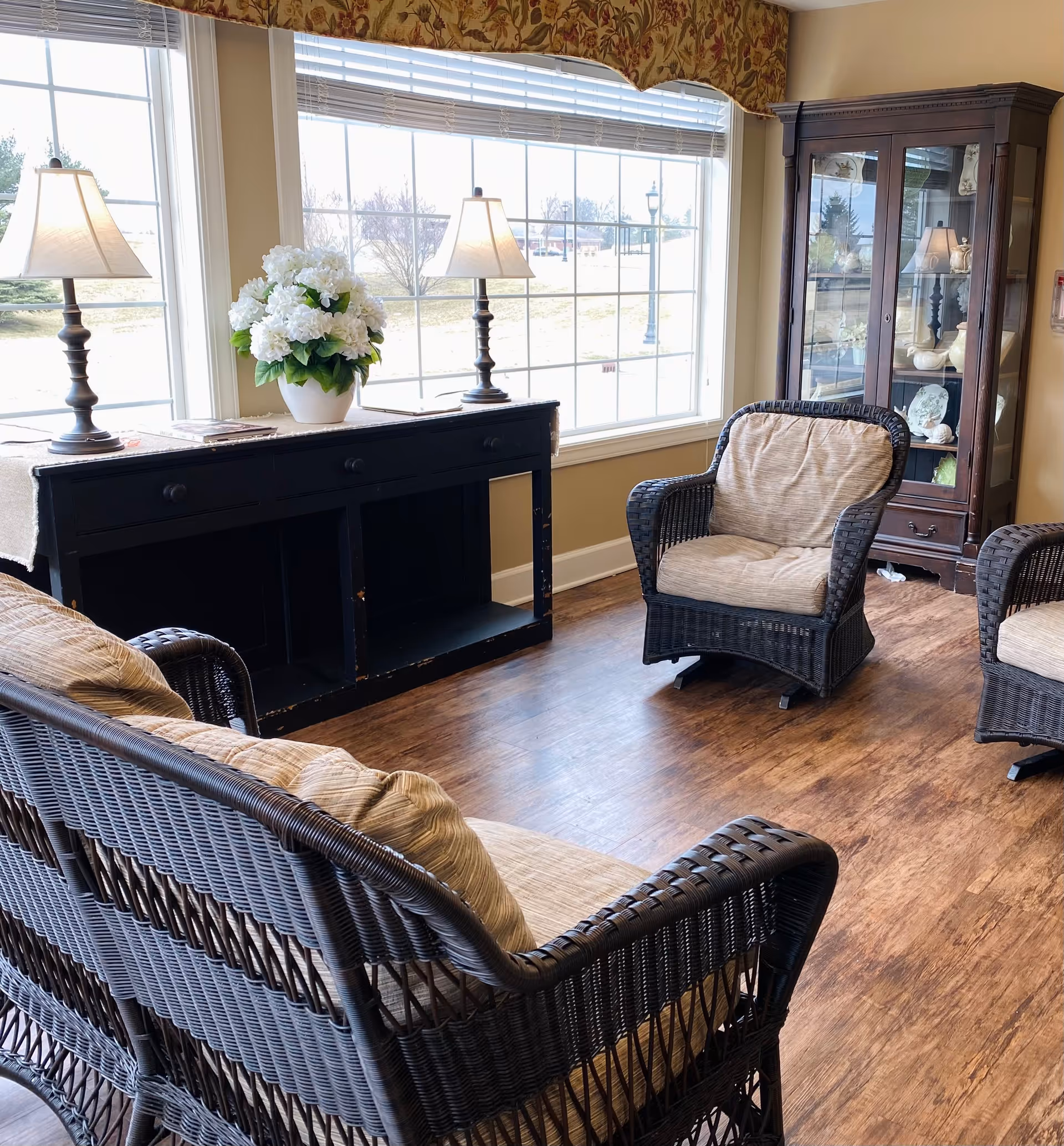 A cozy sitting area with wicker furniture including a loveseat and two armchairs with beige cushions. There is a black console table with two lamps and a white flower arrangement in front of large windows letting in natural light. A wooden display cabinet with decorative items is in the corner of the room. The floor is wood and the walls are painted a warm beige color.