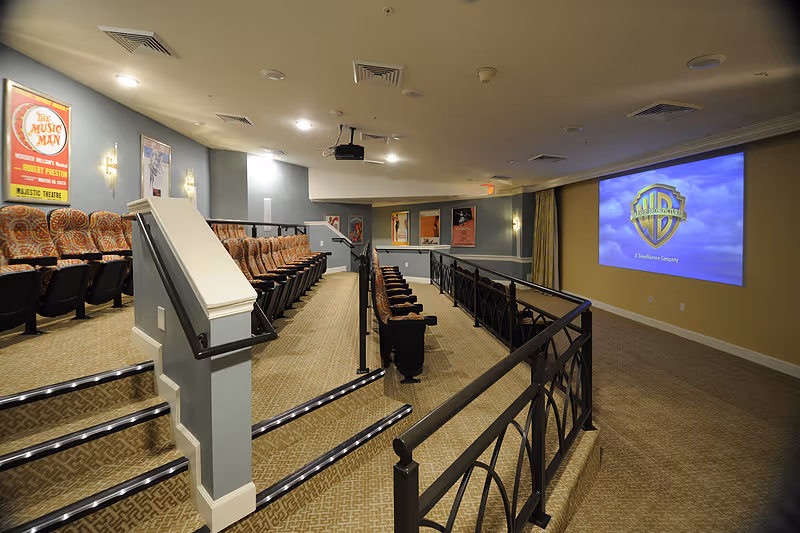 Interior view of a senior living facility's small theater room with tiered seating featuring patterned upholstered chairs. The room has a large screen displaying the Warner Bros. logo on the right wall, movie posters on the left wall, and carpeted floors with handrails along the steps.