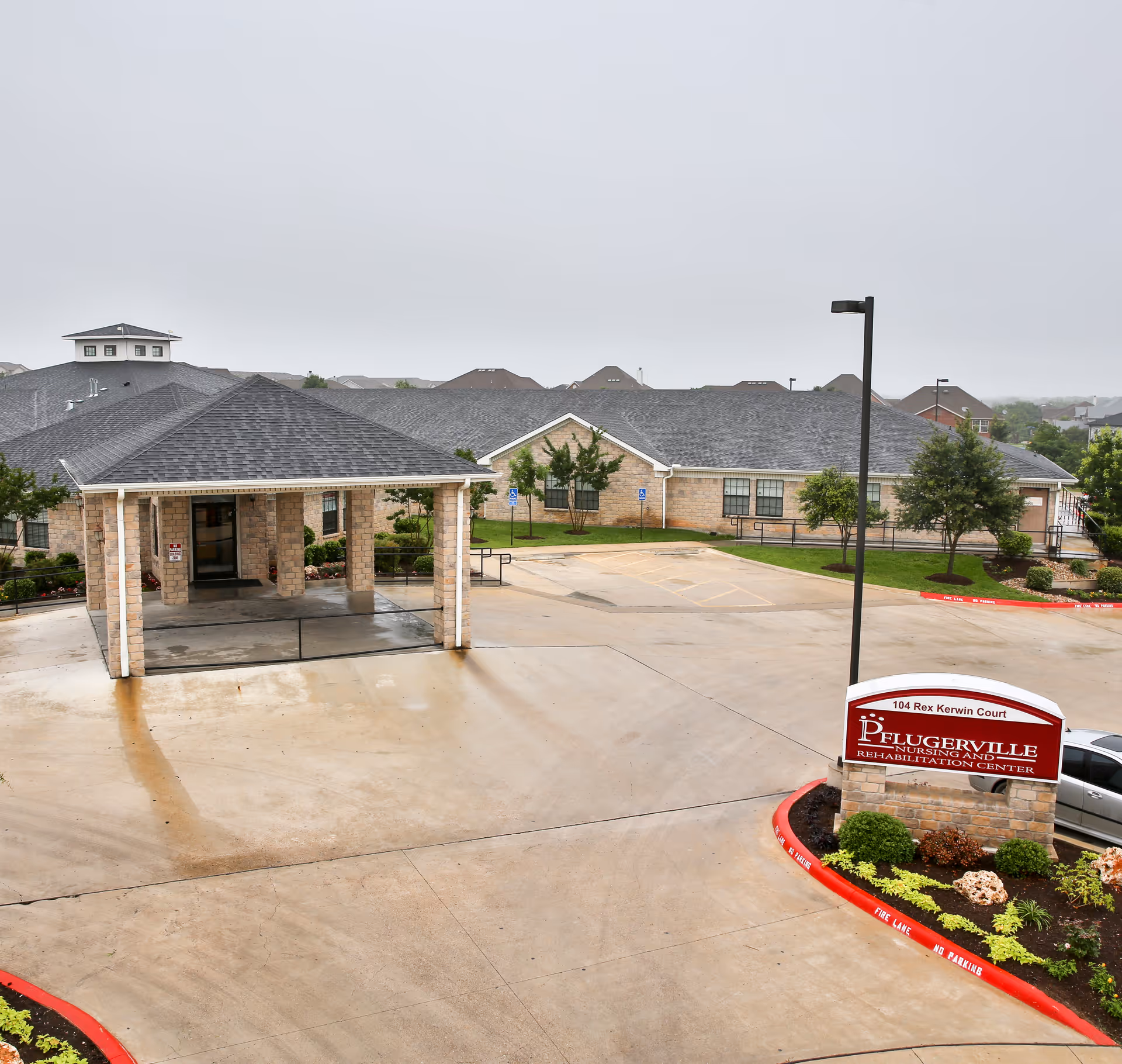 Exterior view of Pflugerville Nursing and Rehabilitation Center showing the main entrance with a covered drop-off area, parking spaces, landscaped greenery, and a sign with the facility's name and address.