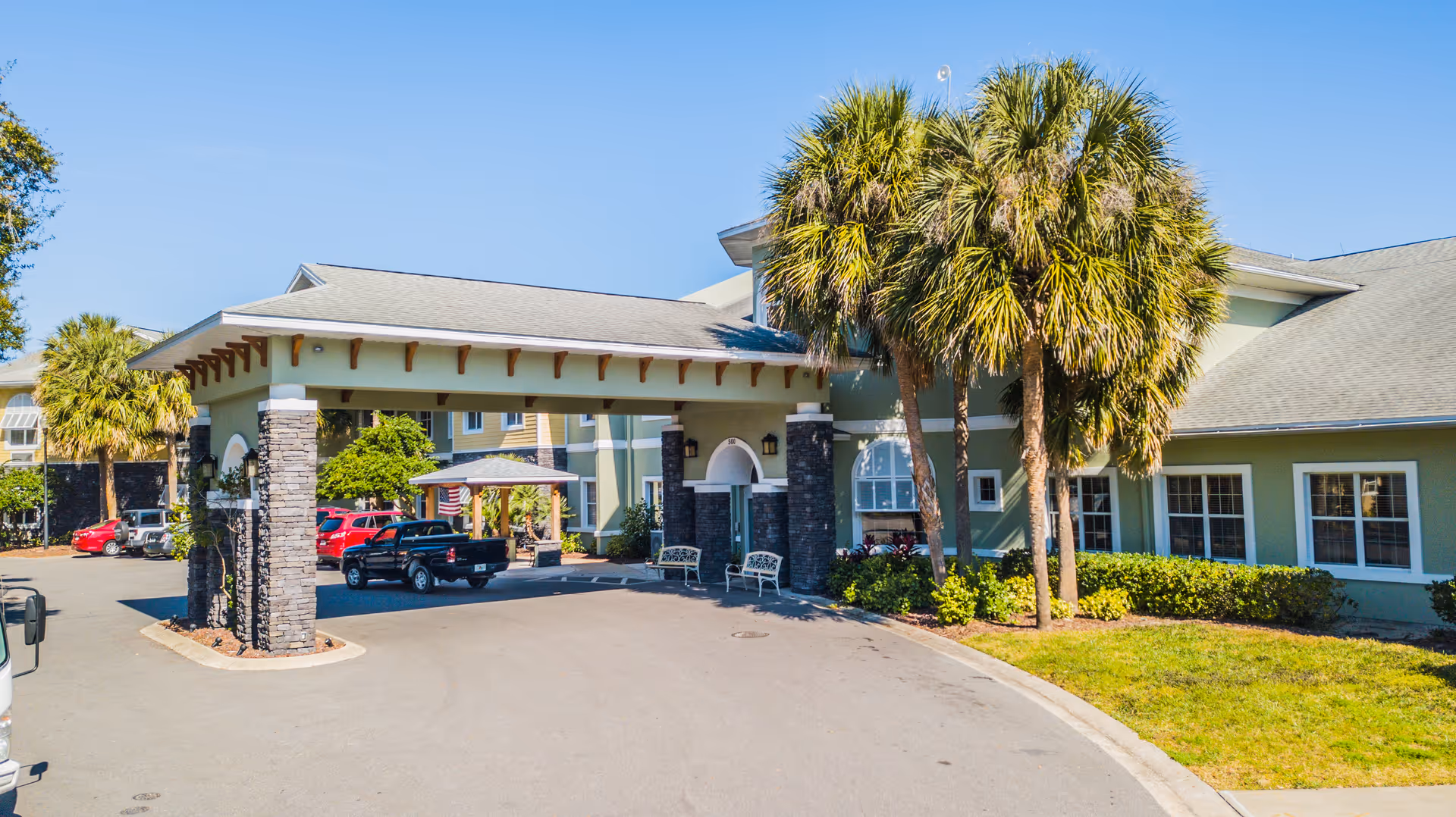 Front exterior view of Bridgewater at Waterman Village Assisted Living facility with a covered entrance, palm trees, parked vehicles, and clear blue sky.