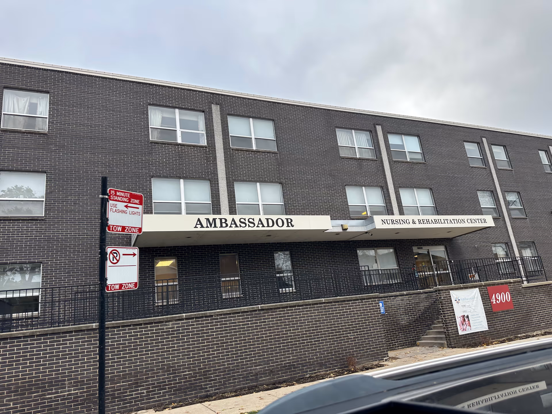 Exterior view of a three-story brick building with a sign reading 'AMBASSADOR NURSING & REHABILITATION CENTER' above the entrance. There are multiple windows and a sidewalk in front with a street sign indicating a 15 minute standing zone and no parking tow zone. The sky is overcast.