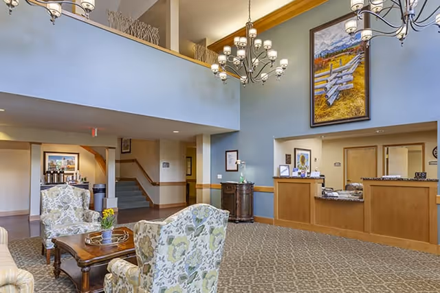 A spacious senior living facility lobby with high ceilings and a chandelier. The room features patterned armchairs around a wooden coffee table with a small plant on it. There is a reception desk on the right side with framed pictures and a large painting of a rustic fence on the wall above it. Stairs and a hallway are visible in the background.