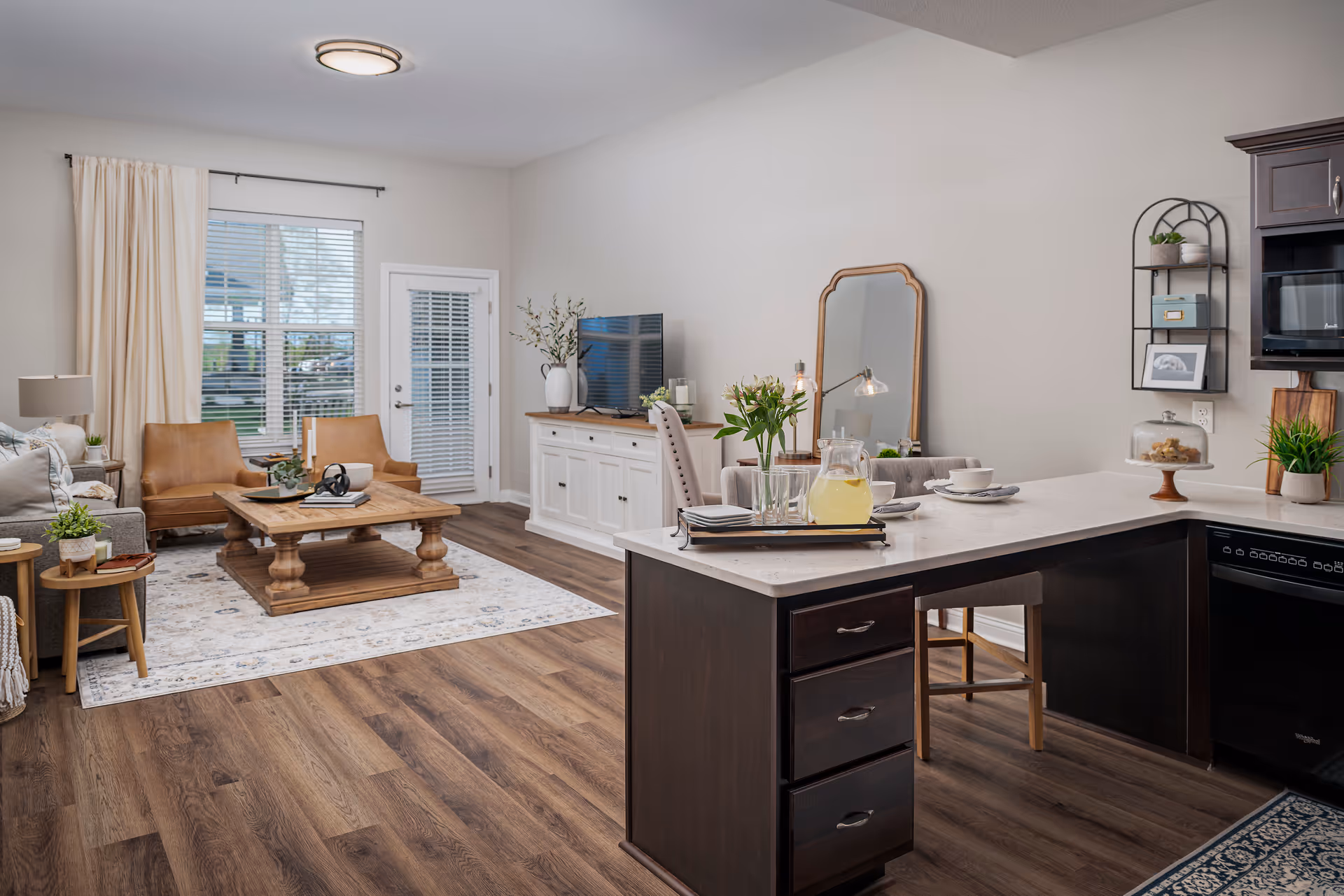 A bright and spacious living area with a modern open floor plan. The room features a cozy seating area with a gray sofa, two tan leather chairs, and a wooden coffee table on a patterned rug. Large windows with white curtains allow natural light to fill the space. Adjacent to the living area is a kitchen with dark wood cabinetry, a white countertop island with bar stools, and various decorative items including plants and a pitcher of lemonade. A white sideboard with a TV and a large mirror on the wall complete the room.
