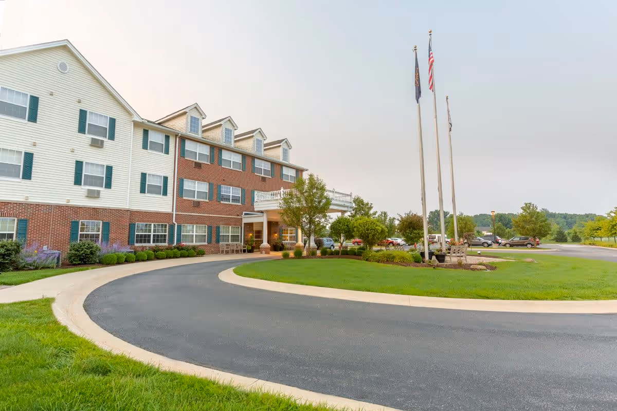 Exterior view of a multi-story senior living facility with a curved driveway, manicured lawn, three flagpoles with flags, and several parked cars in the background under a cloudy sky.