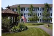 A multi-story white building with multiple windows, surrounded by green grass, trees, and a paved walkway. In the foreground, there is a wooden gazebo and an American flag on a flagpole.