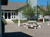 Outdoor patio area at Sonoma Hills Retirement Living Community featuring round concrete picnic tables with attached benches, surrounded by small trees and shrubs, adjacent to a building with multiple windows and a glass door entrance.