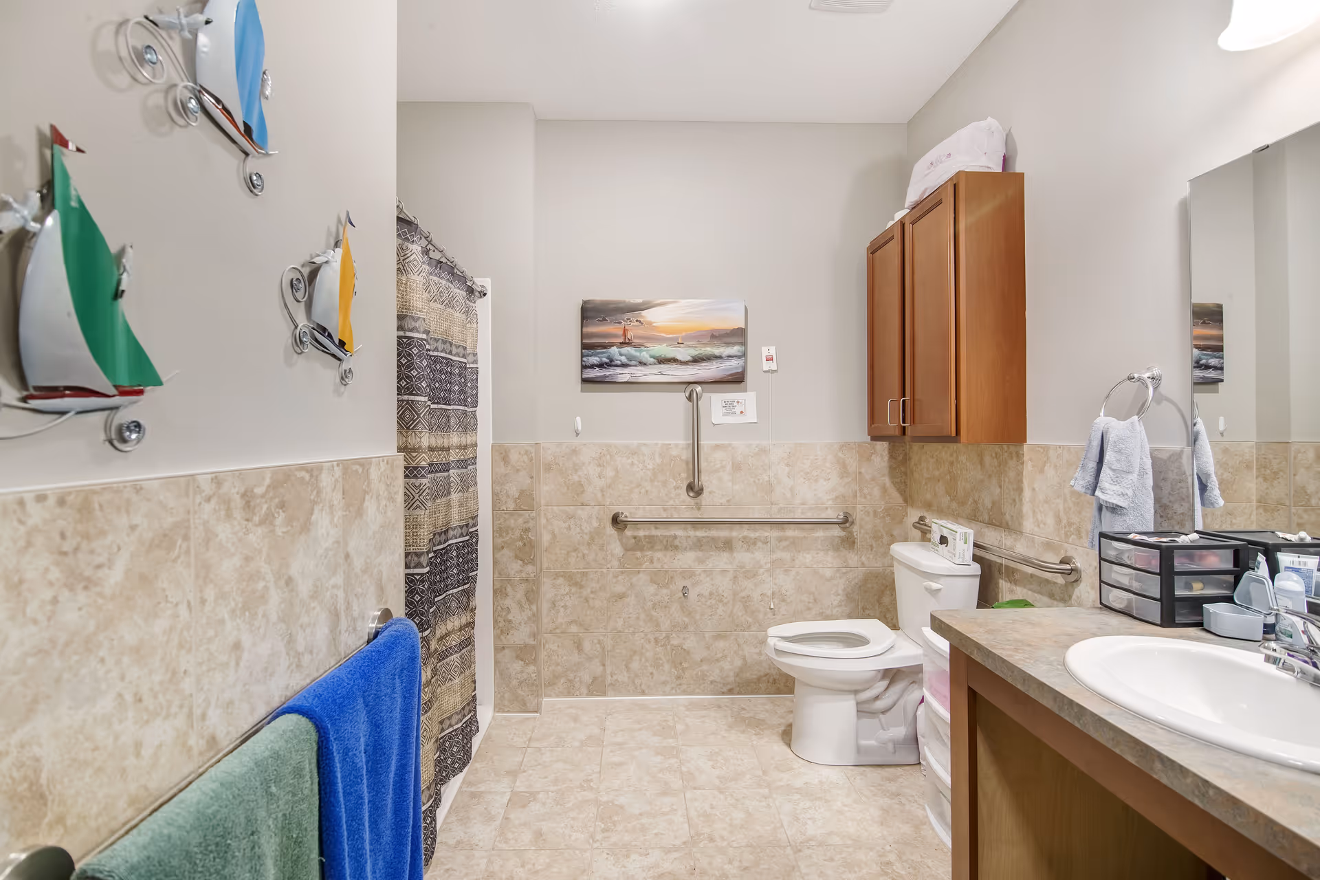 A clean and accessible bathroom with beige tiled walls and floor. The bathroom features a white toilet with grab bars on the walls for support, a shower with a patterned curtain, a wooden cabinet mounted on the wall, and a sink with a countertop and mirror. Colorful metal sailboat decorations are mounted on the wall, and blue and green towels hang on a towel rack.