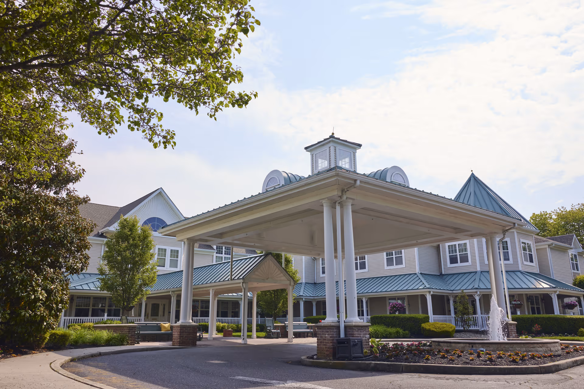 Front exterior view of Brandywine Brandall Estates by Monarch, showing a large covered entrance with white columns, a fountain, landscaped flower beds, and a multi-story building with green metal roofing and numerous windows under a partly cloudy sky.