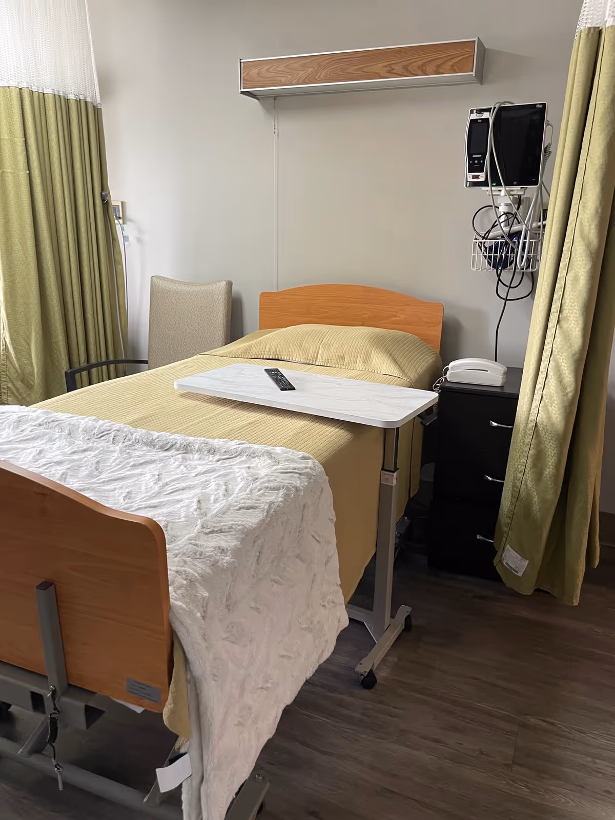 A hospital-style bed with a yellow blanket and a white textured throw blanket at the foot. A small overbed table with a remote control rests on the bed. To the right of the bed is a black bedside cabinet with a white telephone and medical monitoring equipment mounted on the wall above it. Green curtains partially frame the bed on both sides, and a beige chair is positioned near the head of the bed against a plain light-colored wall.