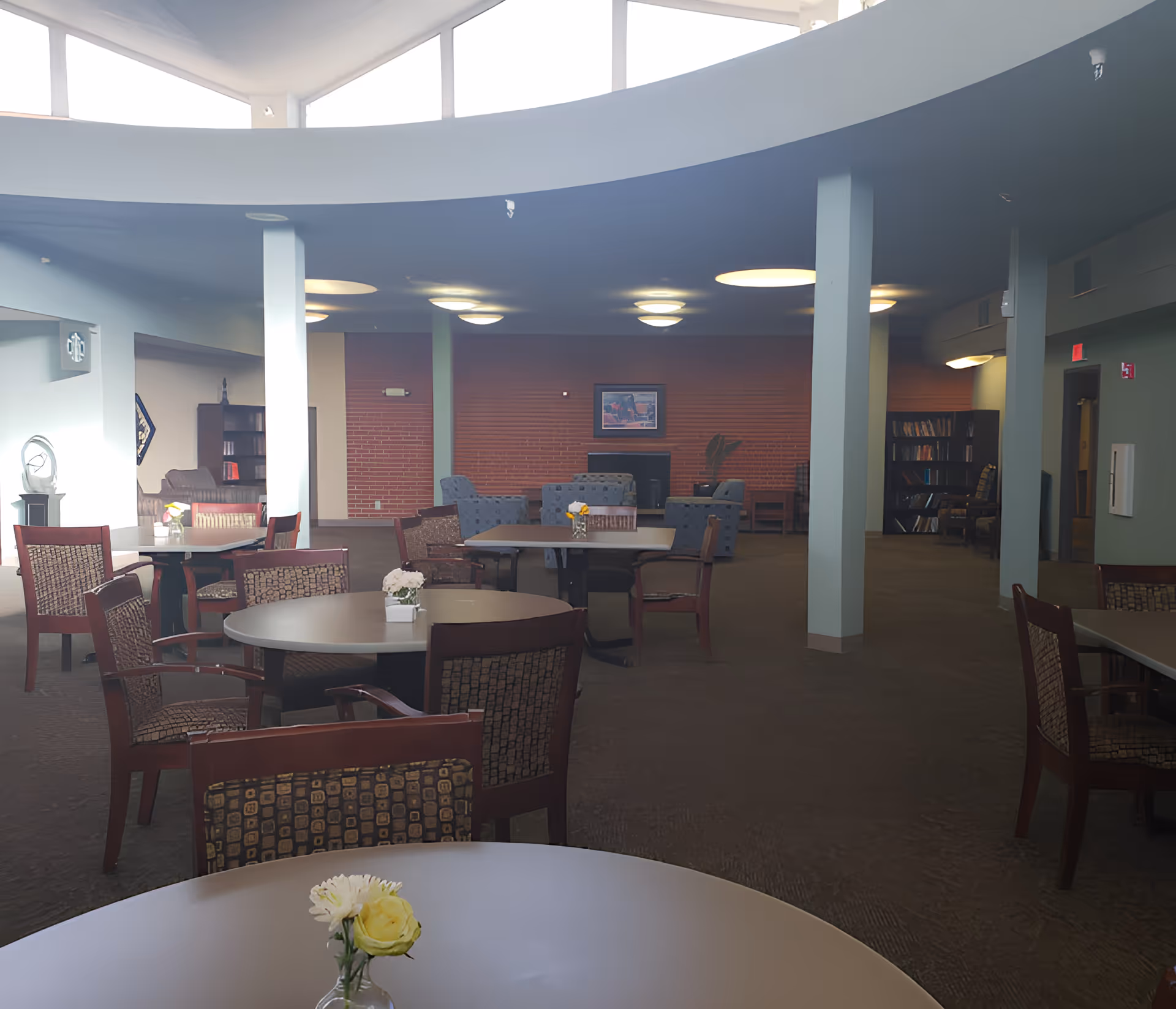 A spacious common area with round tables and chairs arranged for seating. The room features a high ceiling with large windows letting in natural light. In the background, there are comfortable armchairs, bookshelves, and a brick accent wall with a framed picture. The space appears to be a lounge or living room area in a senior living facility.