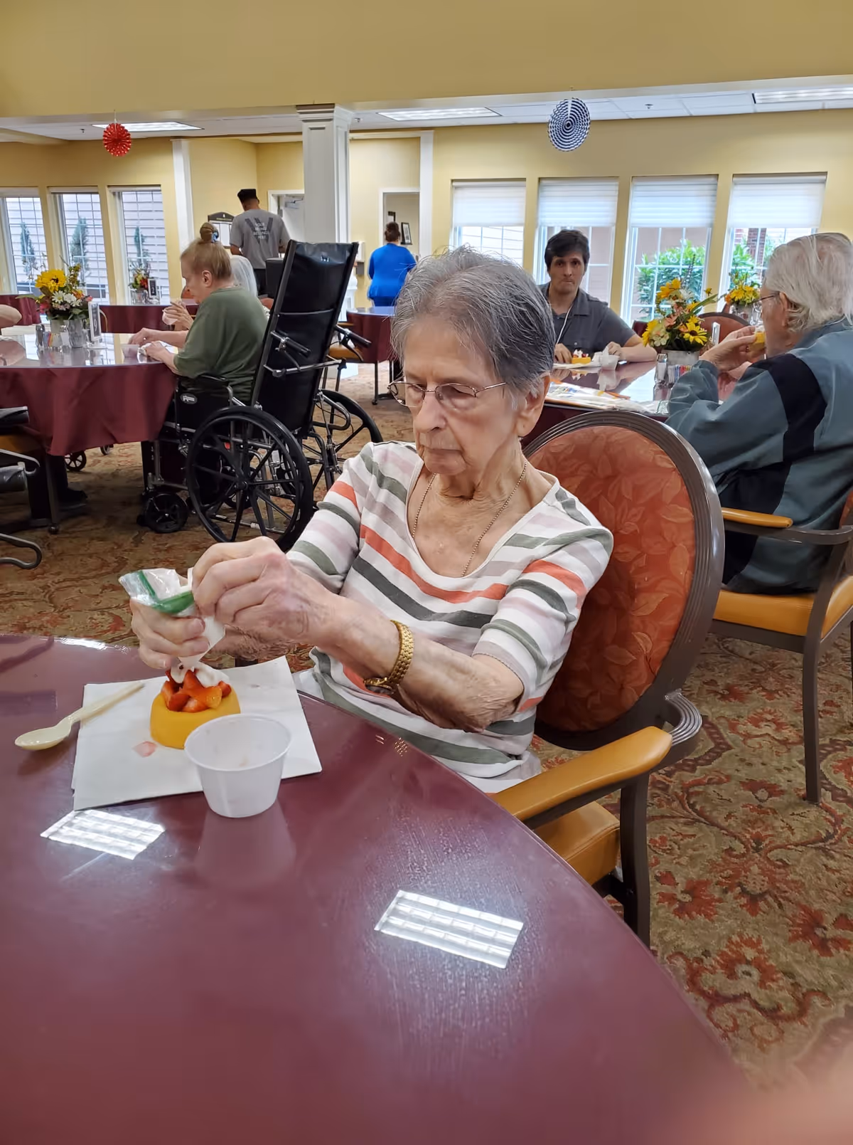 An elderly woman sitting at a round table in a dining area, decorating a small dessert with strawberries. Other elderly individuals and caregivers are visible in the background, some seated and some standing. The room has large windows with blinds and is decorated with flowers on the tables.
