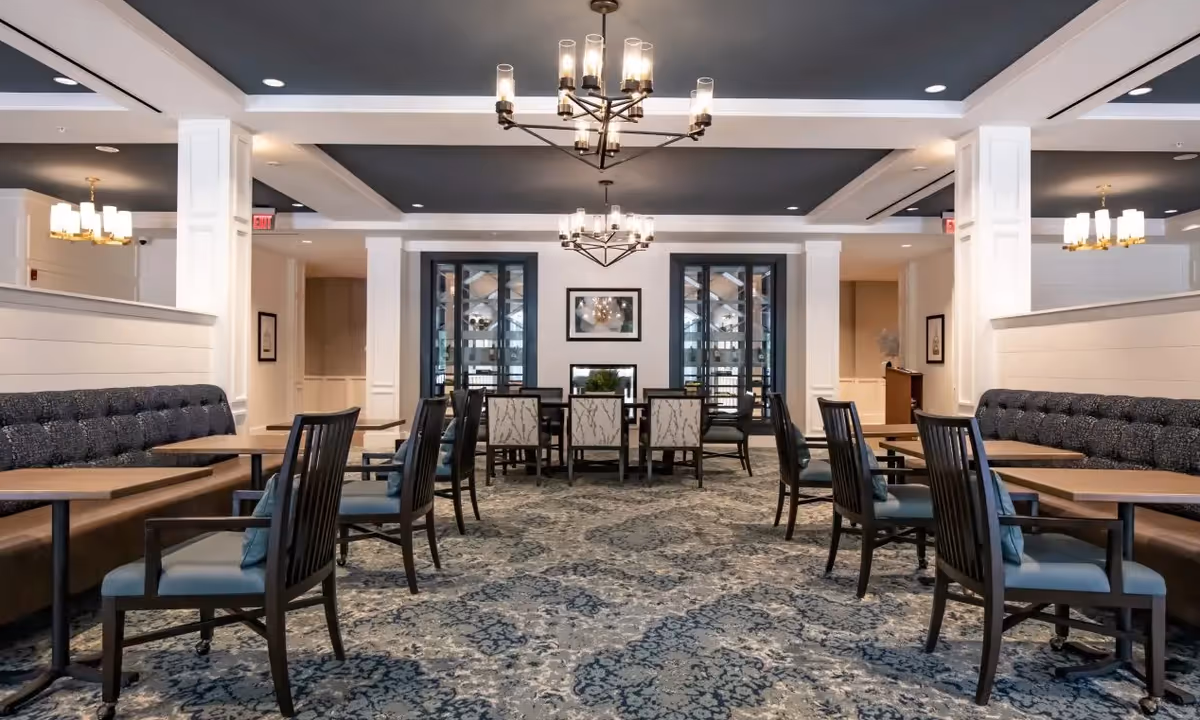 A spacious dining room with multiple wooden tables and chairs arranged neatly. The room features a patterned carpet, cushioned bench seating along the walls, and modern chandeliers hanging from the ceiling. The walls are painted white with decorative molding, and there are framed pictures and glass doors at the far end.