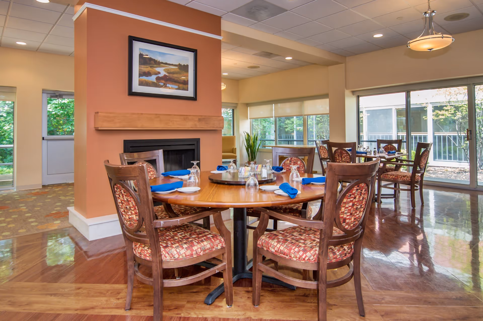 A dining room with round wooden tables set with plates, glasses, and blue napkins. The room features wooden chairs with patterned upholstery, a fireplace with a landscape painting above it, large windows letting in natural light, and a polished wooden floor.