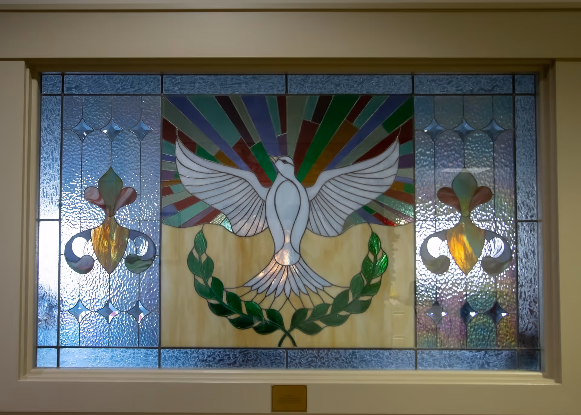 A stained glass window featuring a white dove with outstretched wings above a green laurel wreath, with colorful rays emanating from behind the dove and decorative fleur-de-lis patterns on either side.