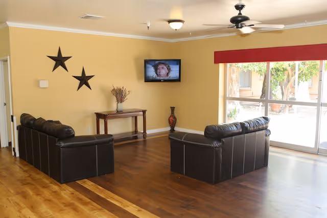 A living room area with two black leather sofas facing a wall-mounted television. The walls are painted yellow, decorated with two black star-shaped wall hangings. A wooden table with a vase of dried flowers and a decorative vase on the floor are placed against the wall. Large windows with a red valance allow natural light to enter the room.