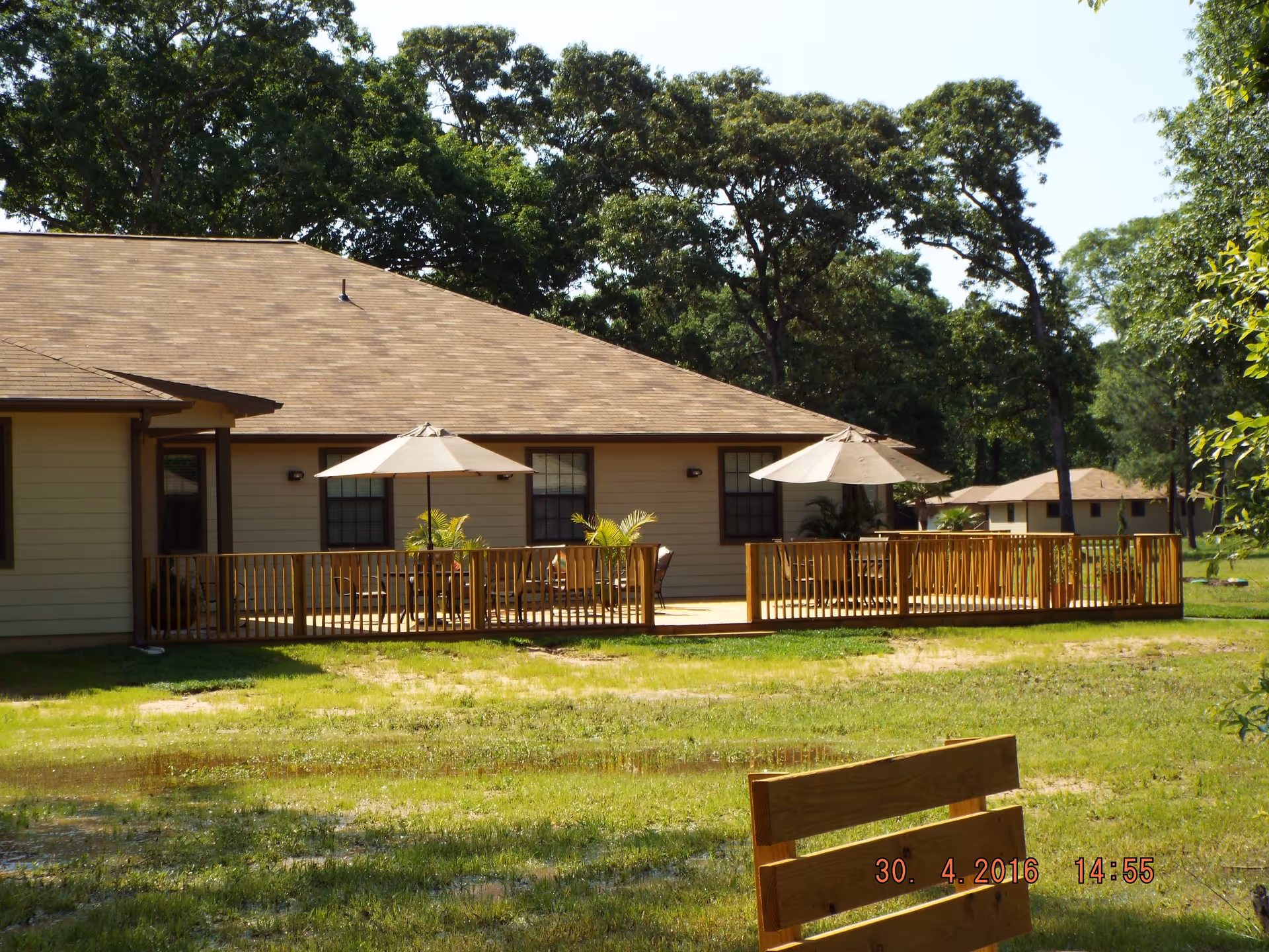 Single-story building with a wooden deck featuring patio umbrellas and chairs, a grassy yard and trees surrounding it.