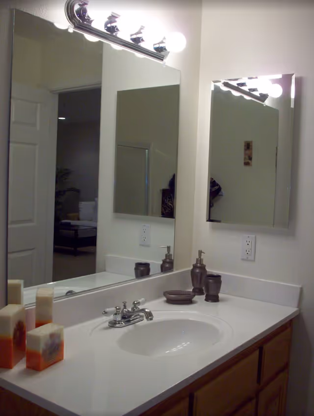 Bathroom sink area with a white countertop, silver faucet, soap dispenser, toothbrush holder, and decorative soap bars. Two mirrors are mounted on the walls above the sink, with a light fixture above the larger mirror. A door is open in the background, showing part of a living room with a couch and table.
