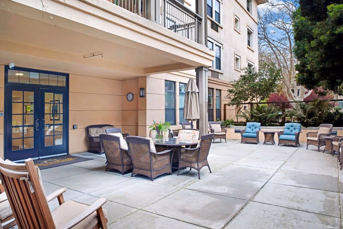 Outdoor courtyard patio with wicker chairs, tables, umbrellas and the building entrance of a senior living facility.
