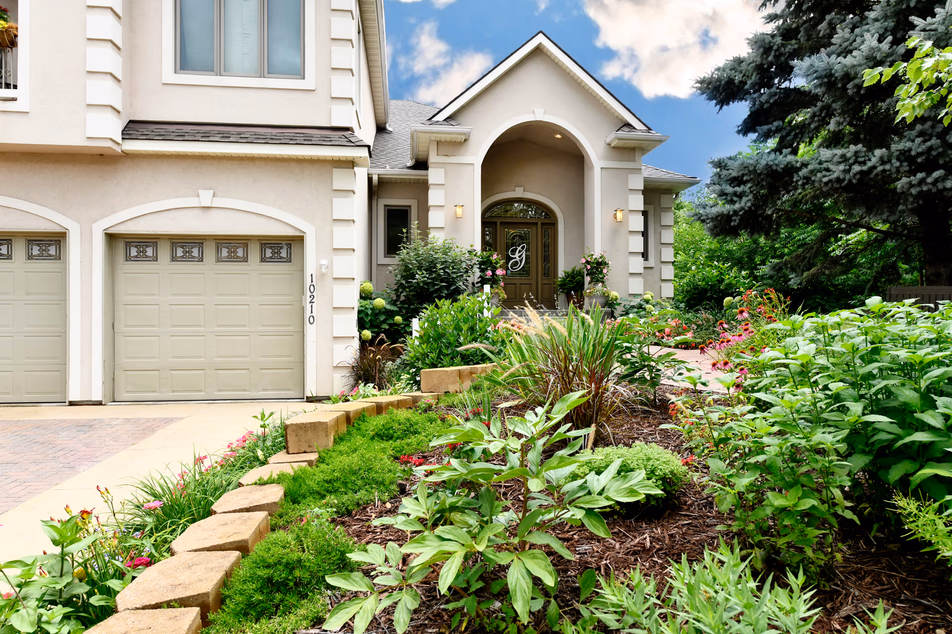 Front exterior view of a residential-style building with a double garage, arched entrance, and well-maintained garden with various green plants and flowers in the foreground under a partly cloudy sky.