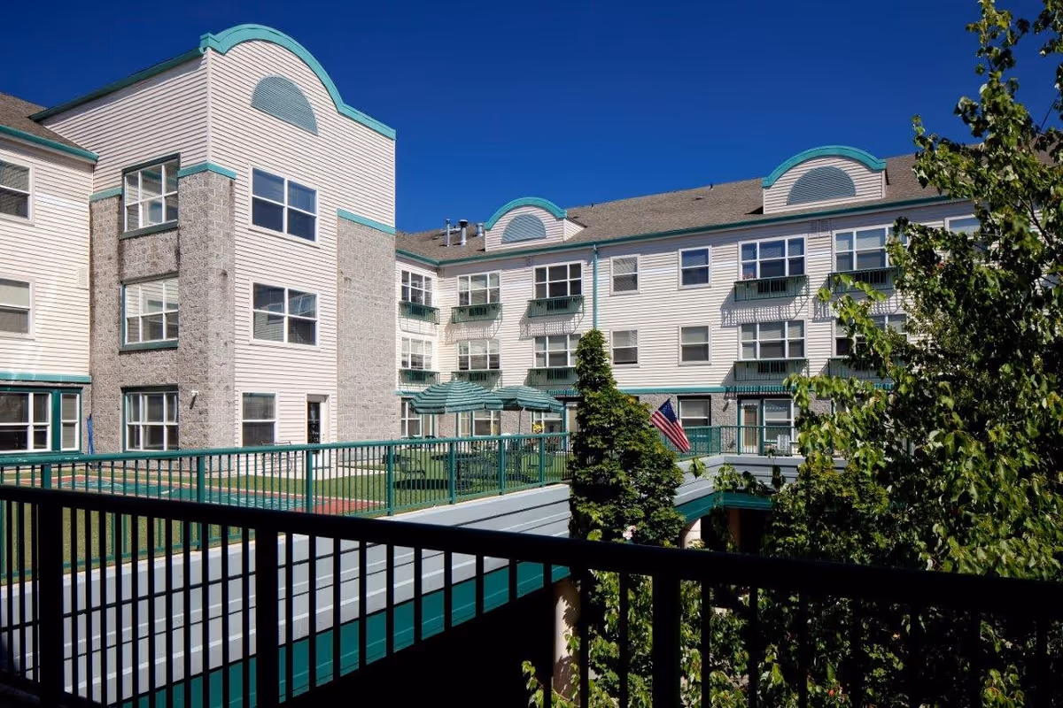 Exterior view of a multi-story residential building with white siding and stone accents, featuring green railings and balconies. There are trees and an American flag visible, with a clear blue sky in the background.