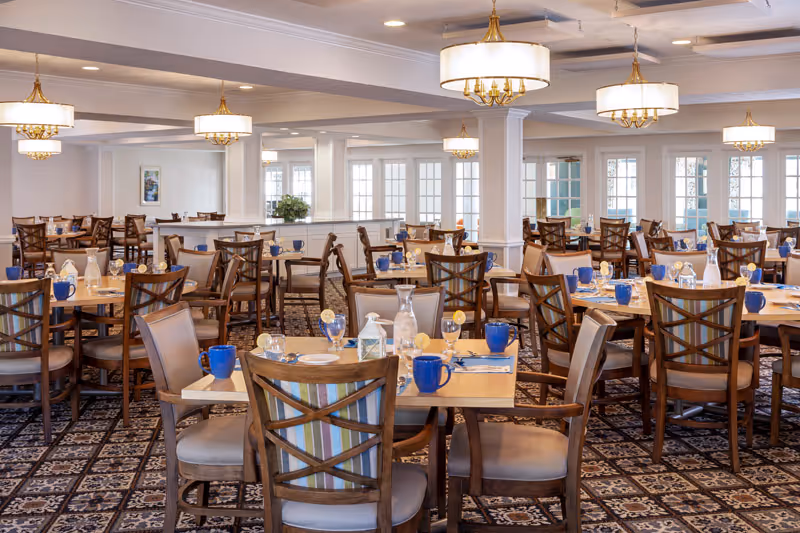 Spacious dining room with multiple set tables and wooden chairs, patterned carpet, and hanging chandeliers.