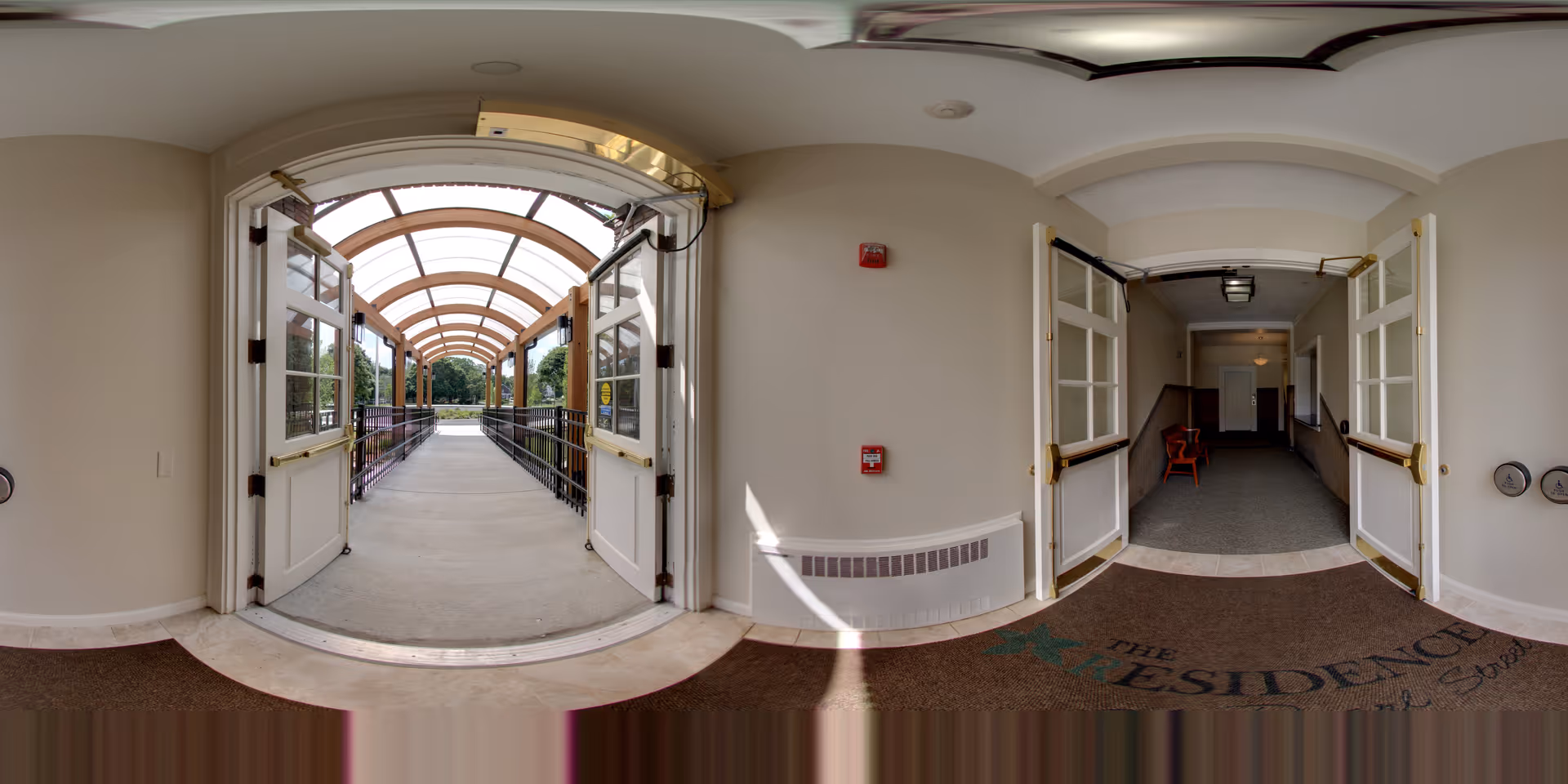 View from inside a building looking out through double doors onto a covered walkway with a curved transparent roof. The walkway leads outside to a green outdoor area. Inside, there is a hallway with open double doors and a red chair visible in the corridor. The floor mat near the entrance has the text 'The Residence at Pearl Street'.