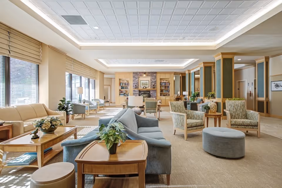 A spacious and well-lit senior living facility common area with multiple seating arrangements including sofas, armchairs, and ottomans. Large windows with blinds allow natural light to fill the room. The back wall features built-in bookshelves and a fireplace with a framed picture above it. The room has a neutral color palette with beige walls, carpeted floors, and wooden furniture accented with plants and decorative items.