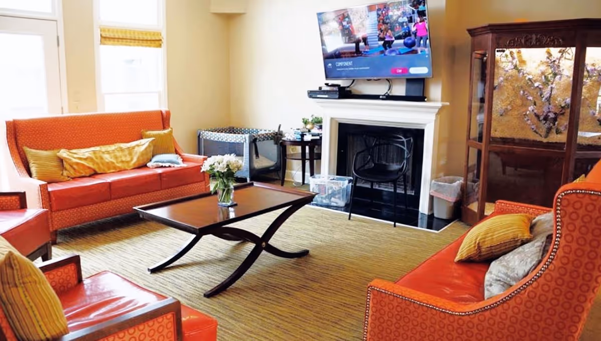 A cozy living room with orange upholstered sofas and chairs arranged around a dark wooden coffee table. A vase with white flowers sits on the table. The room features a fireplace with a black chair in front of it and a flat-screen TV mounted above. There is a large window letting in natural light and a decorative wooden cabinet with a floral design on the right side.