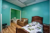 A small bedroom with teal walls and wood flooring. The room contains a single bed with floral bedding, a wooden nightstand, a brown loveseat, and a hospital-style bed in an adjacent area visible through an open doorway. There is also a black rolling stool and a ceiling-mounted air conditioning unit.