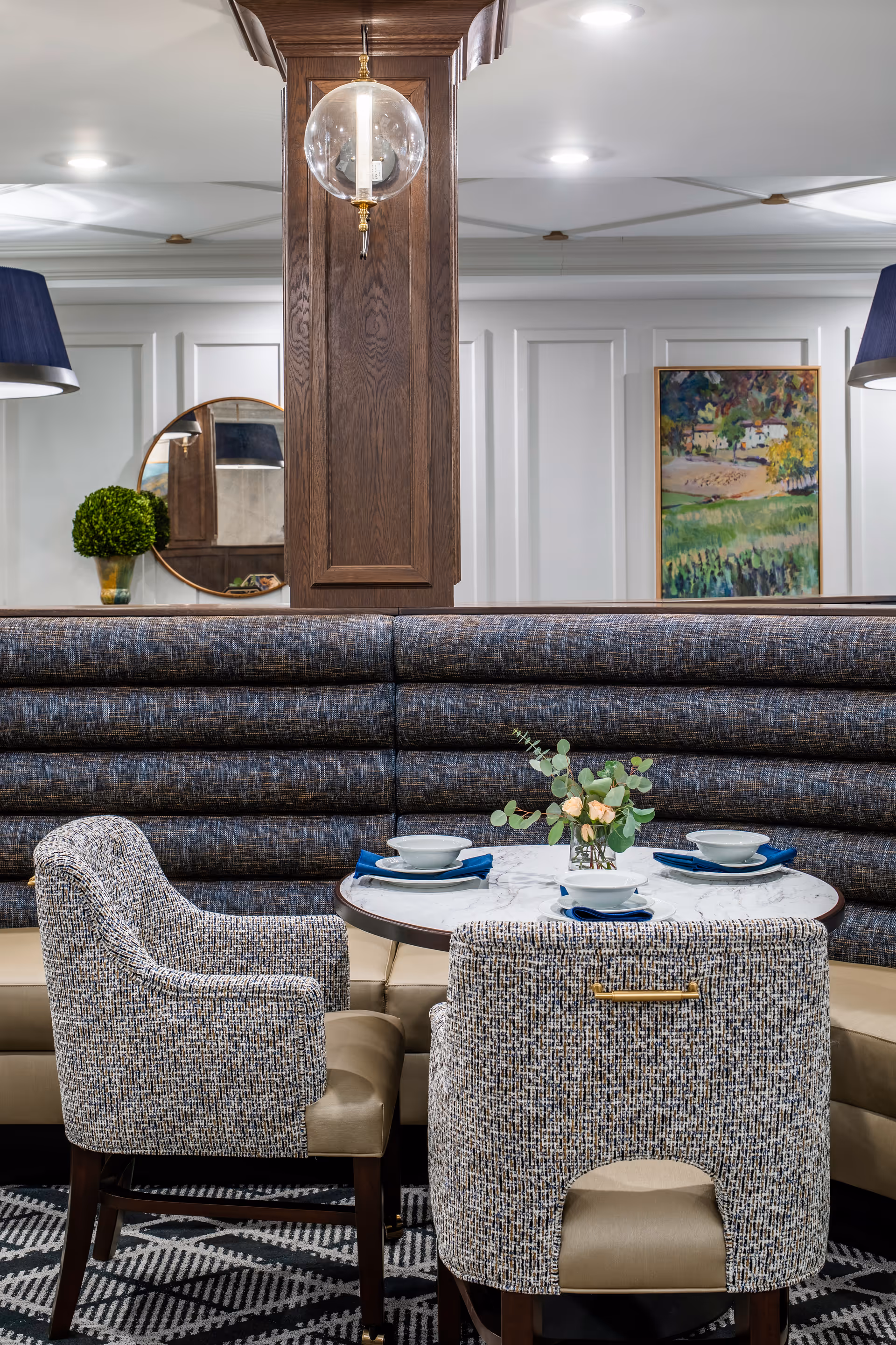 A cozy dining area with a round marble table set with bowls and blue napkins, surrounded by upholstered chairs and a curved banquette with textured fabric. The background features a wooden column with a hanging glass light fixture, a round mirror, a small potted plant, and a colorful framed painting on a white paneled wall.
