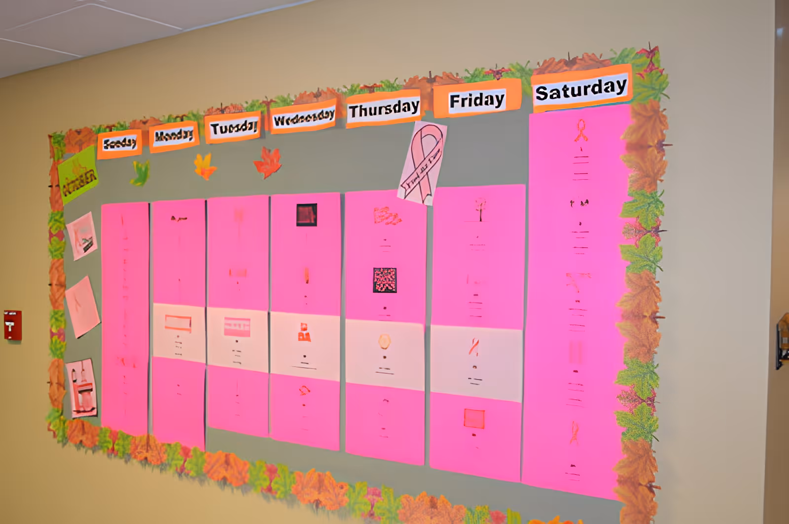 A weekly activity schedule board with days of the week labeled from Sunday to Saturday. Each day has bright pink papers with various activities listed, decorated with autumn leaves around the border and some small decorative cutouts.