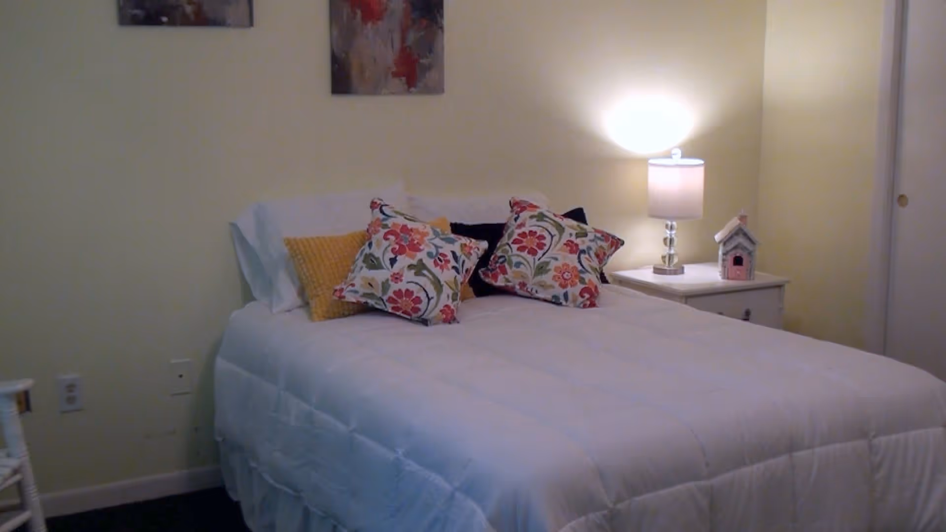 A cozy bedroom with a neatly made bed featuring a white comforter and colorful floral pillows. A bedside table with a lit lamp and a small decorative birdhouse is positioned next to the bed. The walls are painted a soft yellow, and there is a door visible on the right side.
