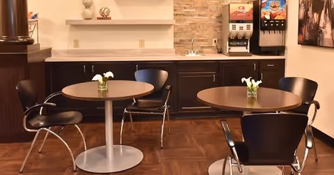 A small dining area with two round tables, each with a small vase of white flowers. Four black chairs with metal legs surround the tables. In the background, there is a countertop with a sink, dark cabinetry, and a beverage dispenser machine. The walls have a light stone tile backsplash and decorative items on a shelf.