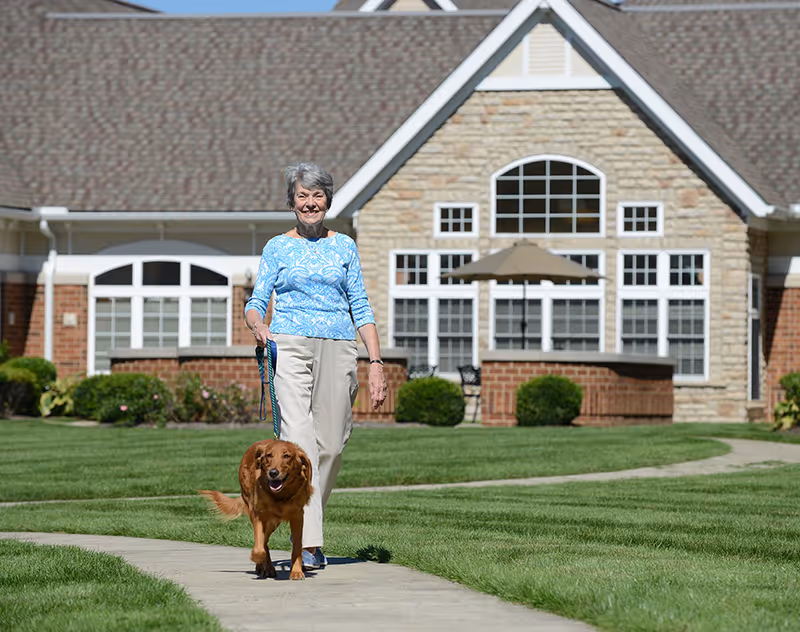 An elderly woman wearing a blue patterned top and beige pants is walking a brown dog on a leash along a paved pathway. Behind them is a building with a stone and brick facade, large windows, and a patio umbrella. The scene is outdoors on a sunny day with green grass and clear blue sky.