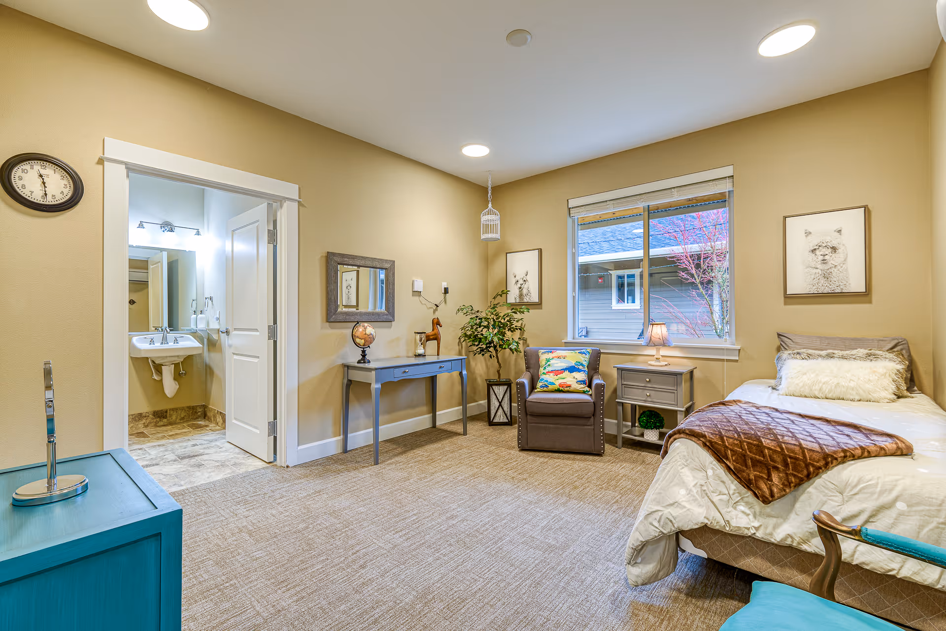 A cozy bedroom in a memory care facility featuring a single bed with white bedding and a brown quilted blanket, a gray armchair with a colorful pillow, a small gray nightstand with a lamp, a blue desk with a globe and decorative items, a window showing an outside view, and an open door leading to a bathroom with a sink and mirror. The walls are beige, and the floor is carpeted.