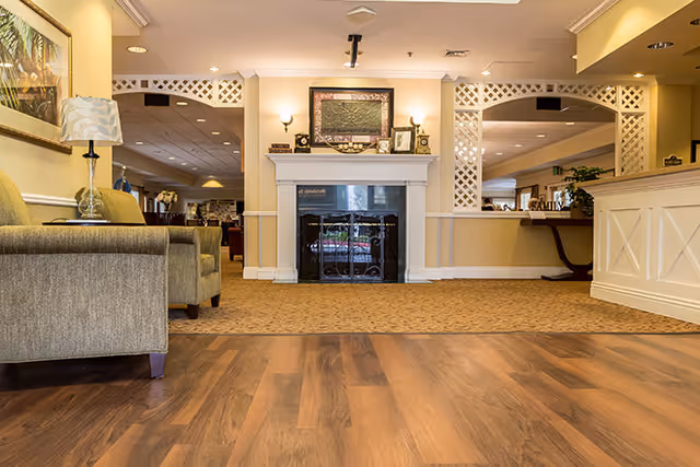 Interior view of a senior living facility lounge area featuring a fireplace with decorative items on the mantel, a beige carpeted floor, wooden flooring in the foreground, a gray armchair with a lamp on a side table, and a reception desk on the right side.
