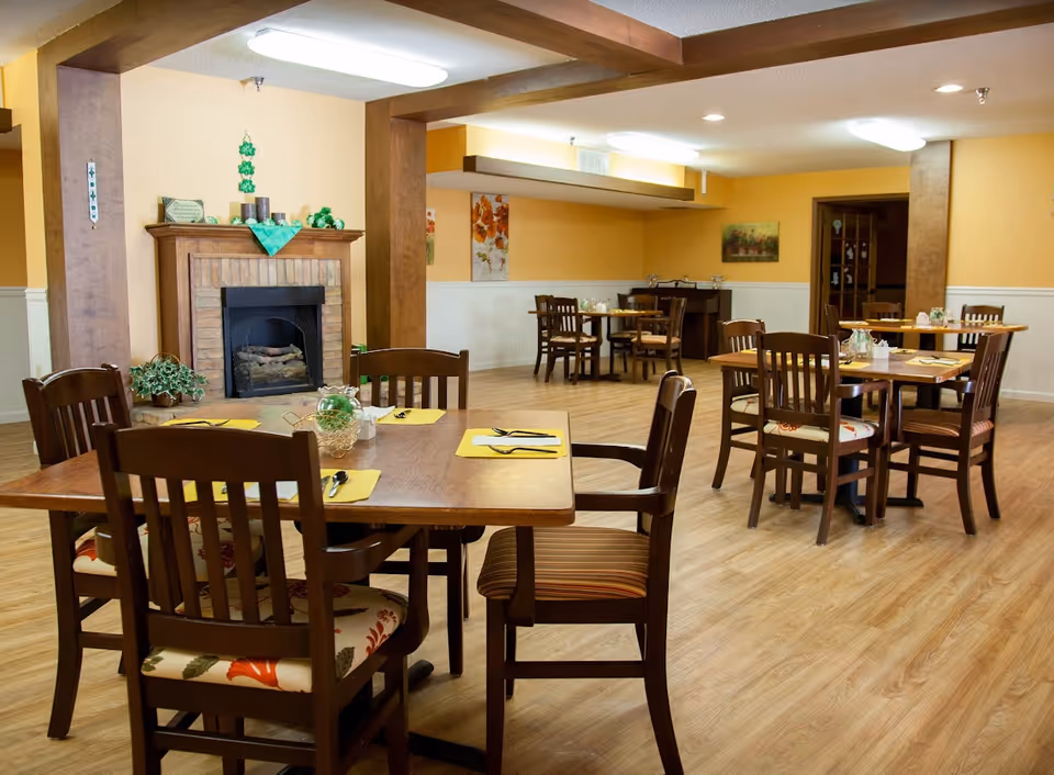 A dining room with several wooden tables and chairs arranged neatly. Each table is set with yellow placemats, utensils, and small decorative centerpieces. The room has light wood flooring, yellow walls with white wainscoting, and a brick fireplace with a wooden mantel decorated with green items. The ceiling has fluorescent lighting, and there are paintings on the walls.