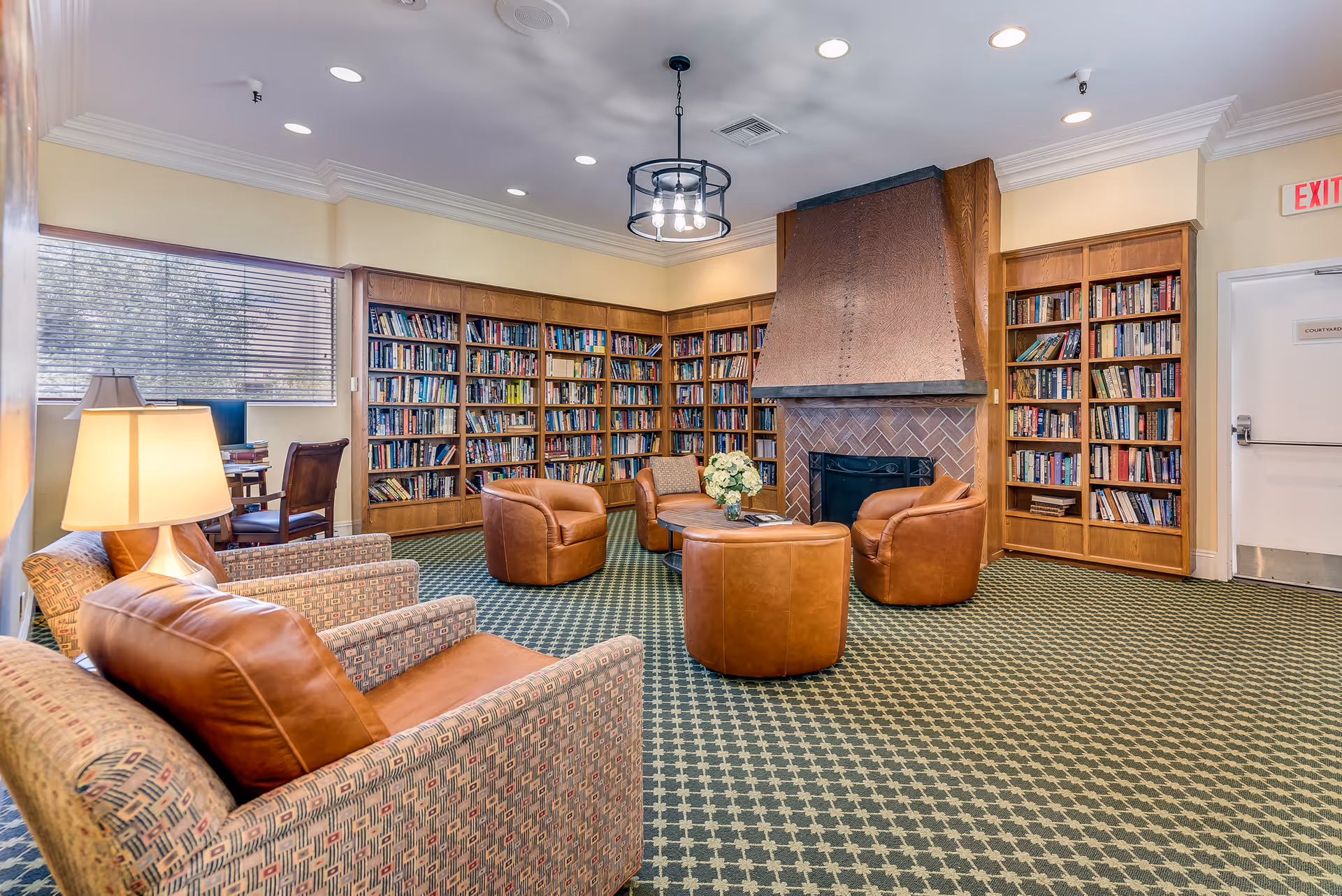 A cozy senior living facility library room with wooden bookshelves filled with books, a large copper fireplace, four brown leather armchairs arranged around a round wooden coffee table with a flower vase, patterned carpet, and a window with blinds letting in natural light.