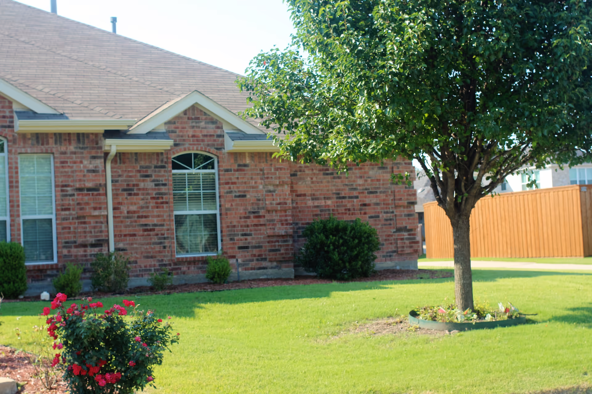 A brick residential building with two windows, surrounded by a well-maintained lawn with a tree and flowering bushes in the foreground. A wooden fence is visible in the background.