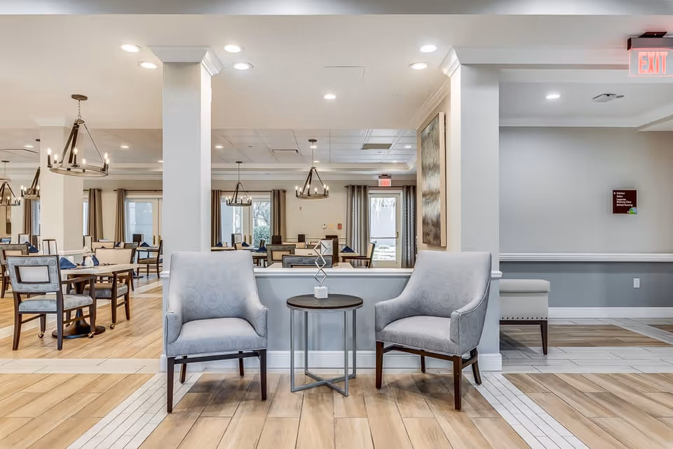 A spacious dining room interior featuring gray upholstered chairs, wooden tables, and modern light fixtures. Large windows allow natural light to fill the room, creating a warm and inviting atmosphere.