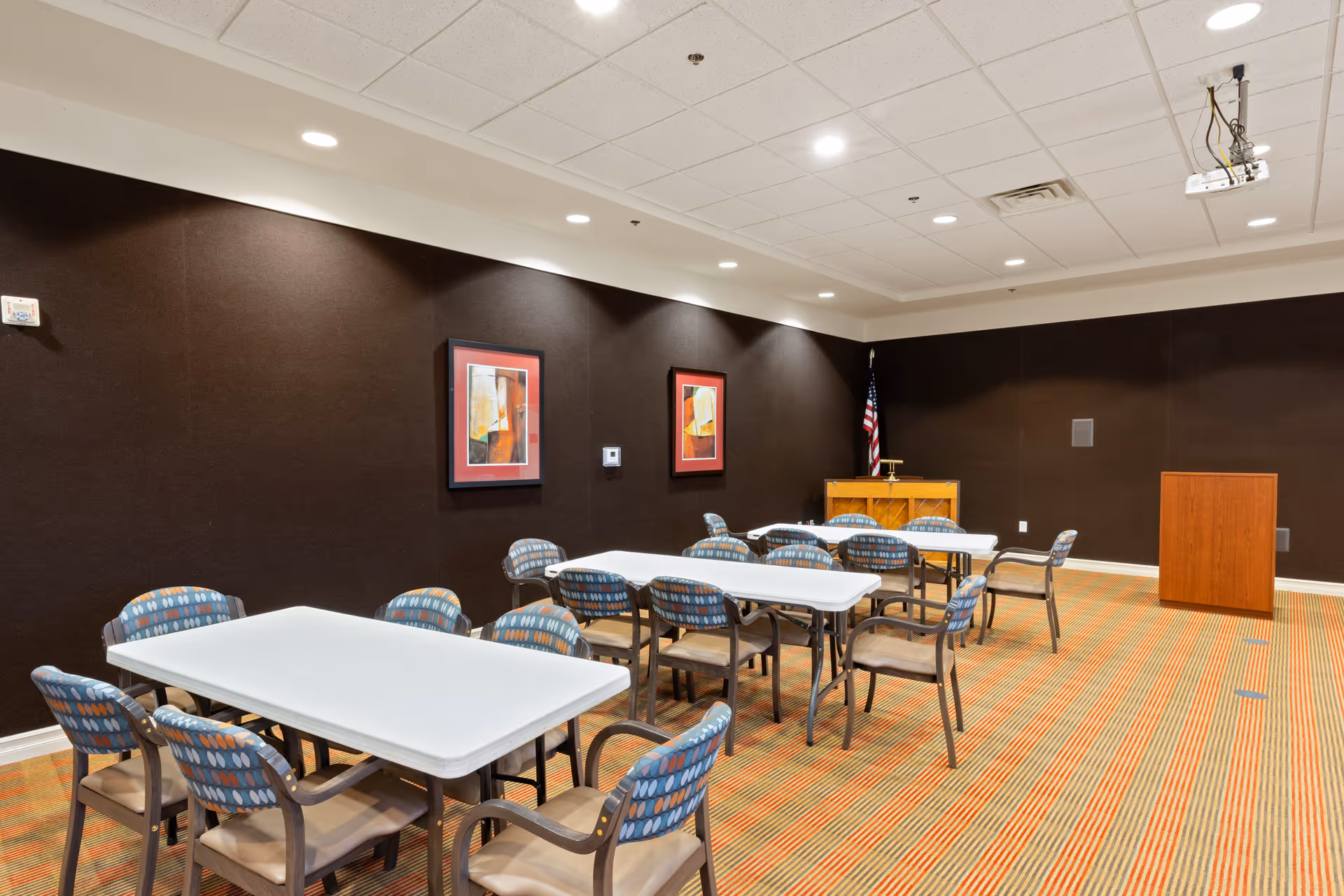 A conference or meeting room with several rectangular white tables and multiple patterned chairs arranged around them. The room has dark brown walls with two abstract paintings framed in red, a wooden podium, an American flag, and a projector mounted on the ceiling. The floor is carpeted with a striped pattern in shades of orange and beige.