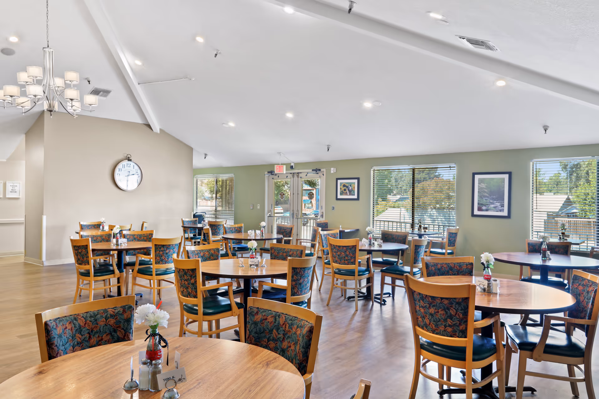 A bright dining room in Almond Grove Assisted Living with multiple round wooden tables and chairs featuring floral upholstery. The room has large windows with blinds letting in natural light, light green walls adorned with framed pictures, a chandelier, and a wall clock. The floor is wooden, and the exit door is visible at the back.