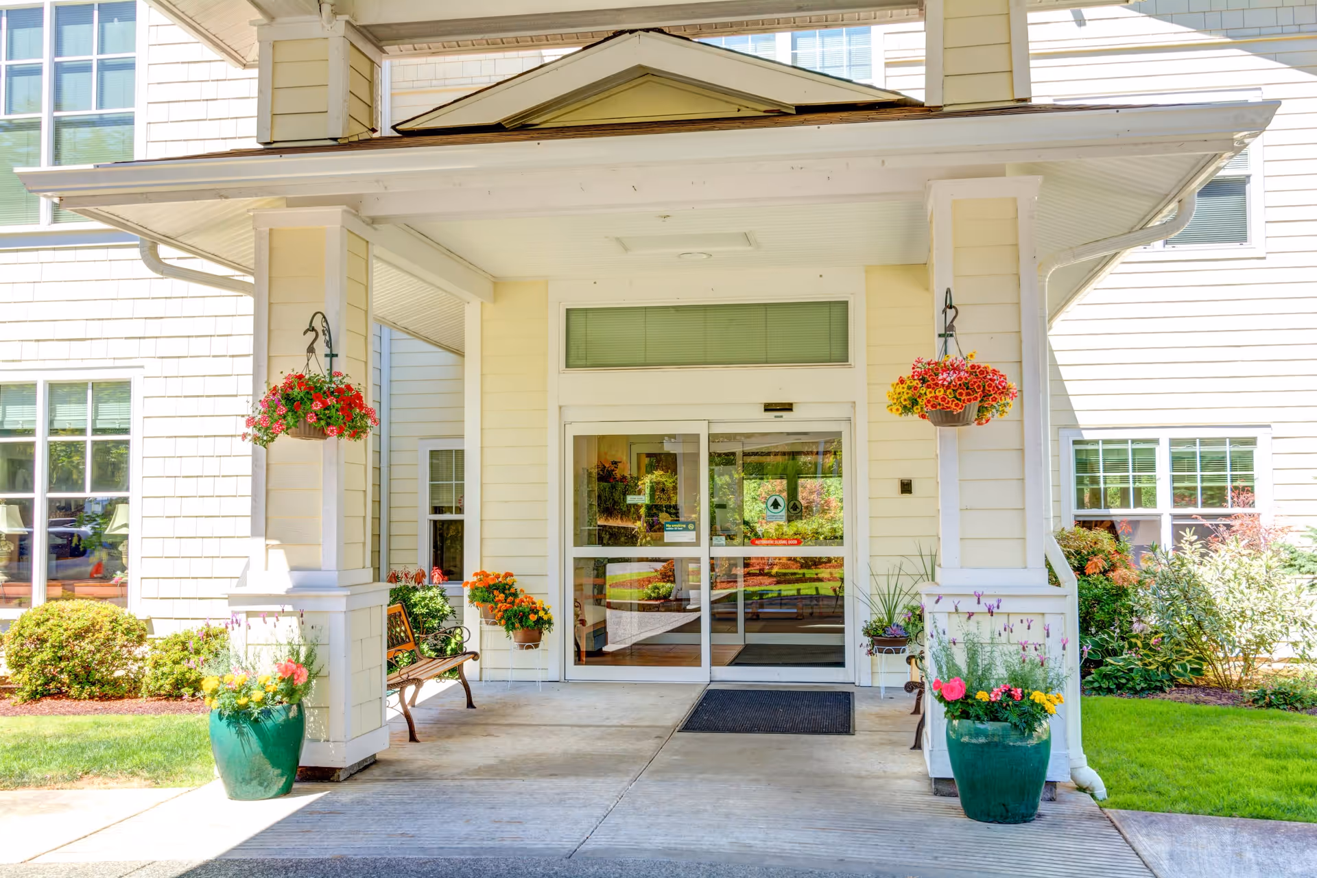 Entrance to a building with a covered porch supported by white columns. There are hanging flower baskets and large green pots with colorful flowers on either side of the entrance. A bench is placed near the door, and the building exterior is light-colored with multiple windows.