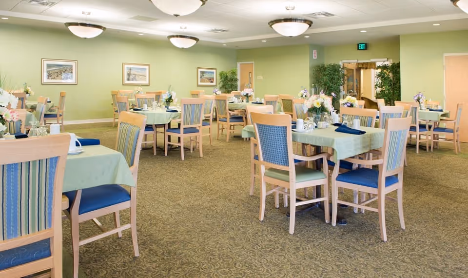 Bright dining room with multiple tables and chairs set with tablecloths and floral centerpieces in a senior living facility.