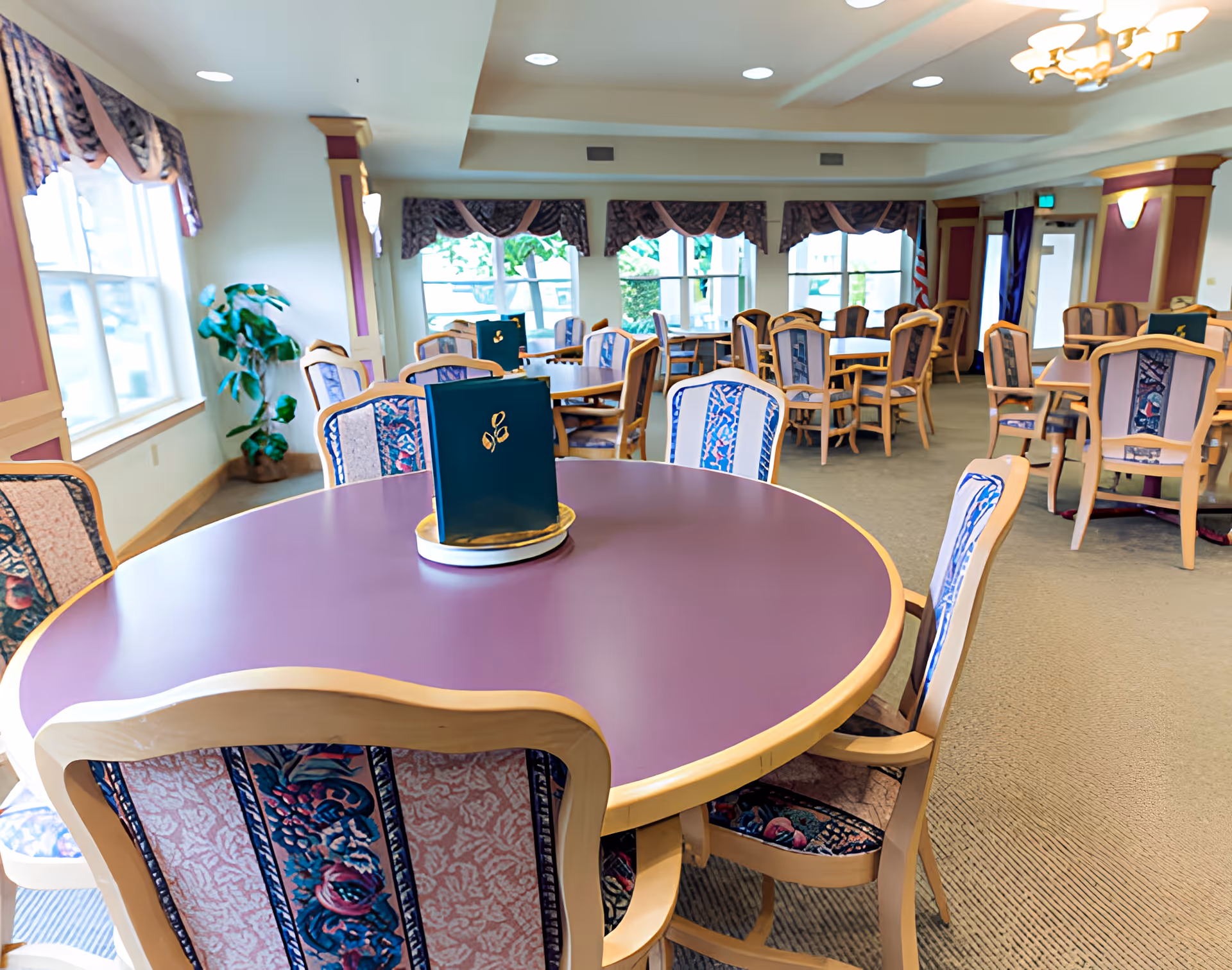 A spacious dining room with multiple round tables and wooden chairs upholstered with patterned fabric. The tables have purple tops and some have green menus placed on them. Large windows with decorative valances allow natural light to fill the room. The walls are decorated with purple and beige accents, and there is a potted plant near one window.